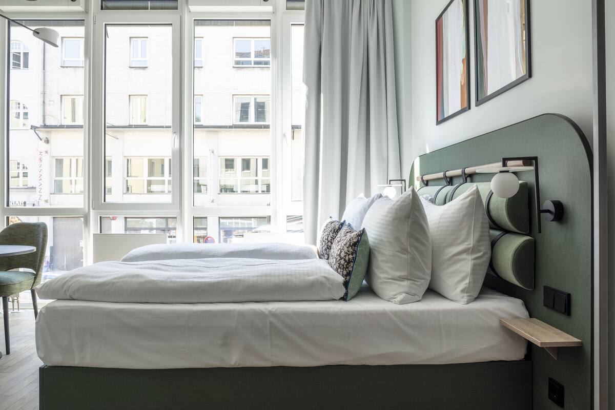 A modern hotel room with a large bed, a bedside table, and a view of the city through large windows.