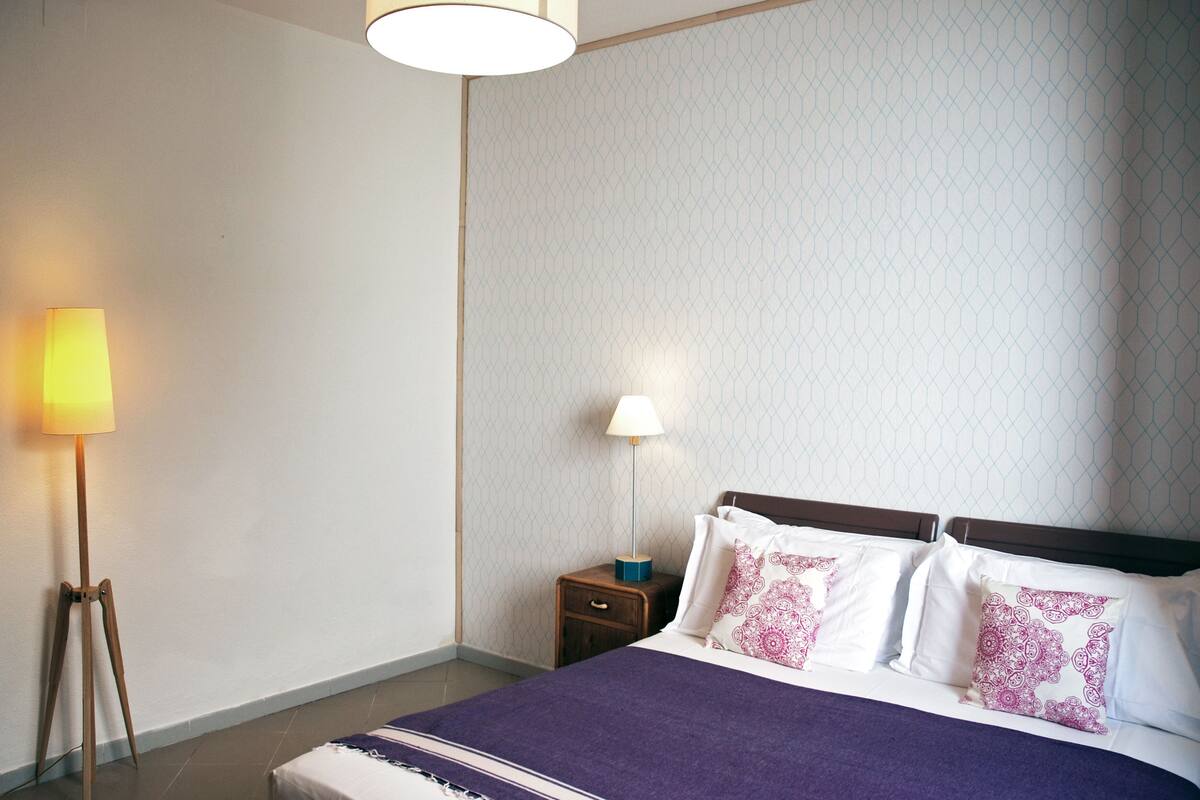 A neatly made bed with a purple blanket, two bedside tables with lamps, and a patterned wall.