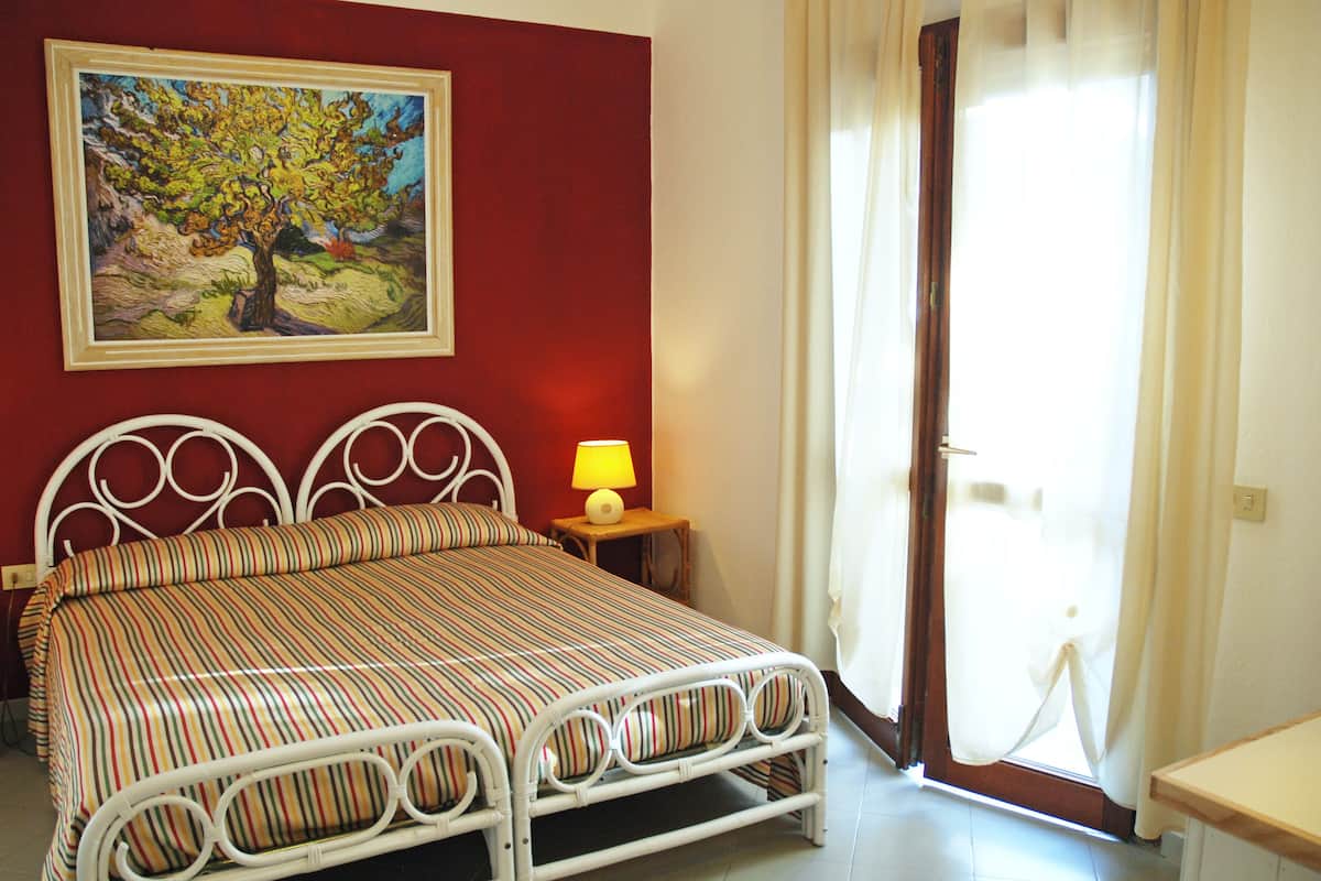 A bed with a striped bedspread, a nightstand with a lamp, a painting on the wall, and a door leading to a bright room.