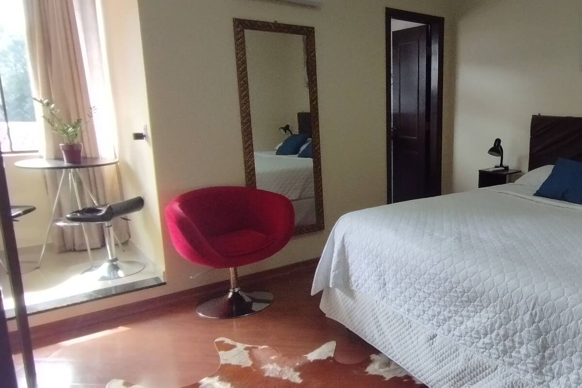 Un dormitorio con una cama grande, una silla roja, una mesita y un espejo.