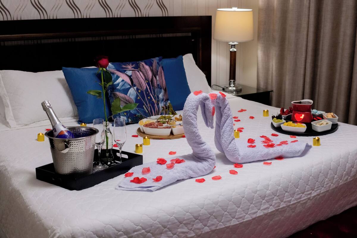 Una cama impecablemente arreglada con un ramo de rosas rojas, una botella de champán y una bandeja de comida y bebidas.
