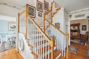 Staircase leads to an upper level, featuring a dining area with a table and chairs, a patterned rug, and framed pictures on the walls.