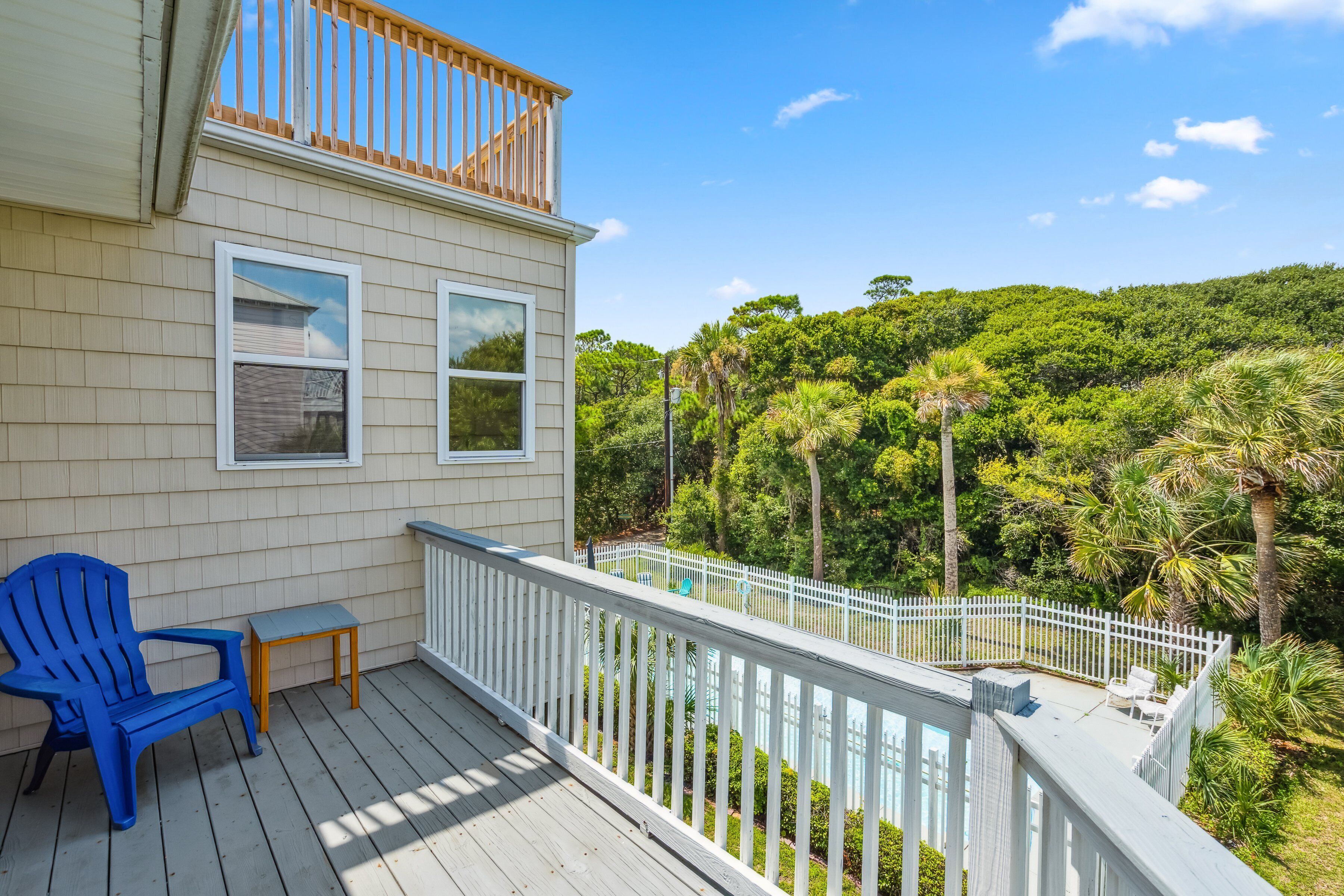 A wooden deck with a blue Adirondack chair and a small table, overlooking a white fence and lush greenery.
