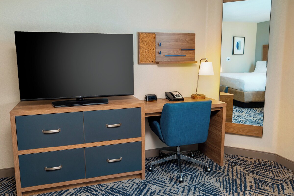 A hotel room with a TV, desk, chair, and a mirror reflecting the bed.