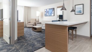 Moderni hotellihuone, jossa on pieni keittiö, sohva, sohvapöytä ja suuri ikkuna.