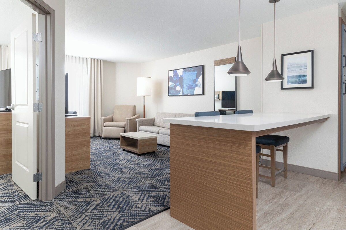 A modern hotel room with a kitchenette, a sofa, a coffee table, and a large window.