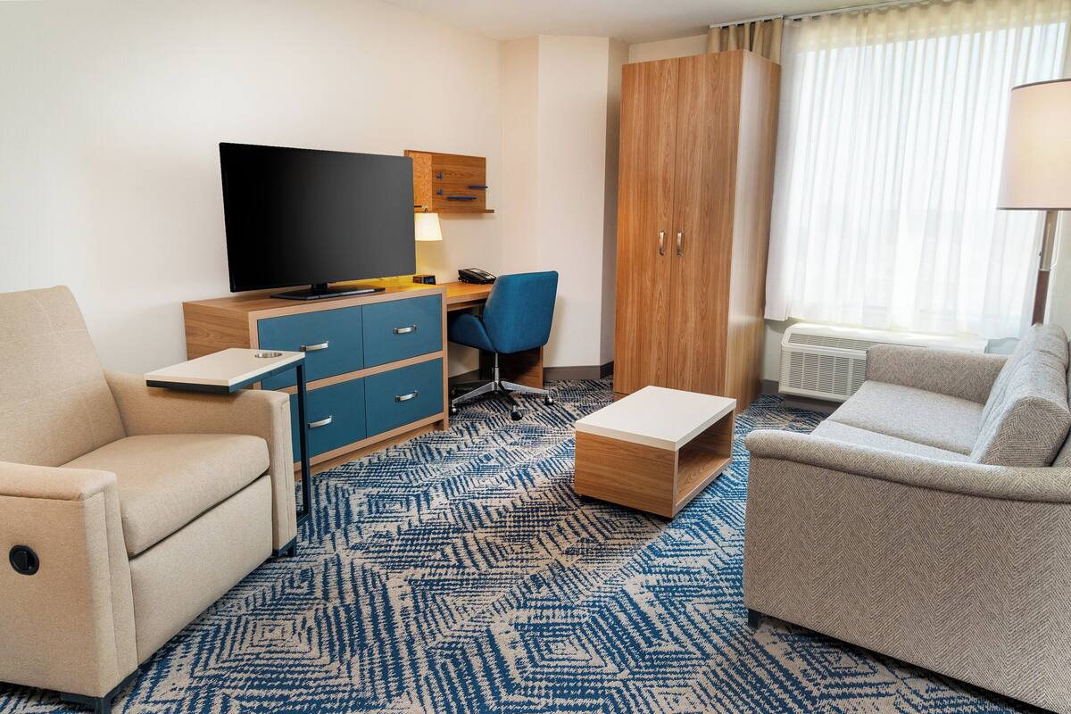 A hotel room with a flat-screen TV, a desk with a chair, two sofas, a wooden wardrobe, and a large window with curtains.