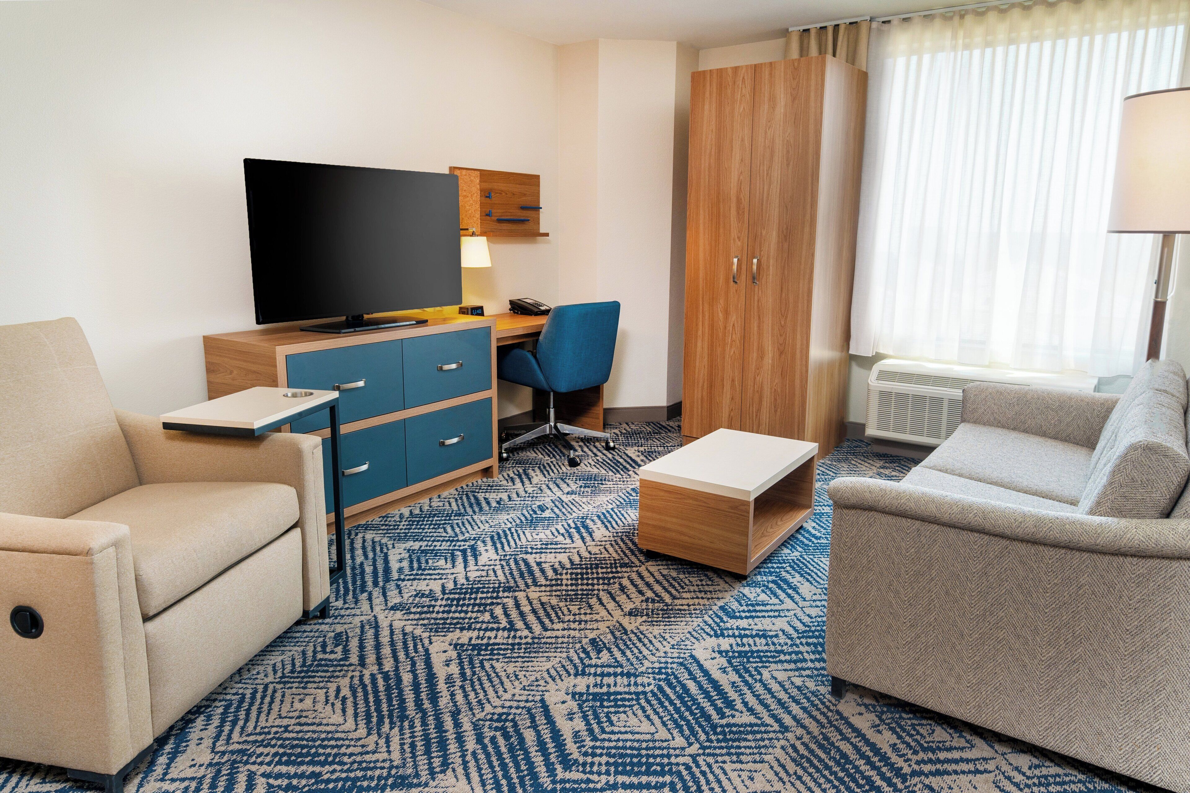 Habitación de hotel con televisor de pantalla plana, escritorio con silla, dos sofás, armario de madera y ventana grande con cortinas.