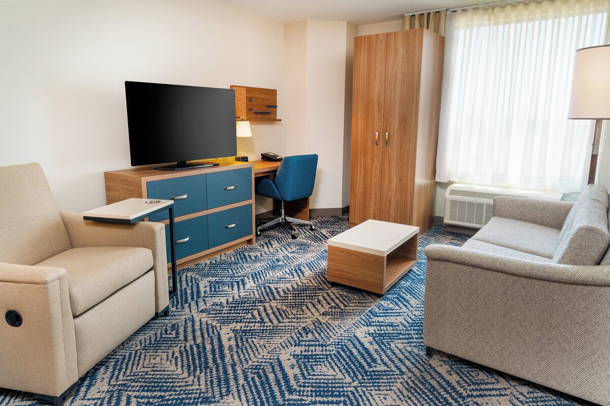 Une chambre d’hôtel équipée d’un téléviseur à écran plat, d’un bureau avec une chaise, de deux canapés, d’une armoire en bois et d’une grande fenêtre avec des rideaux.