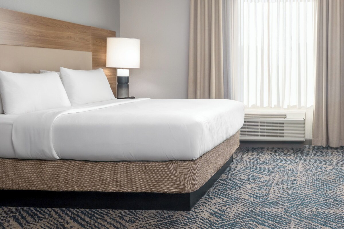 A hotel room with a bed, a nightstand with a lamp, a window with curtains, and a patterned carpet.