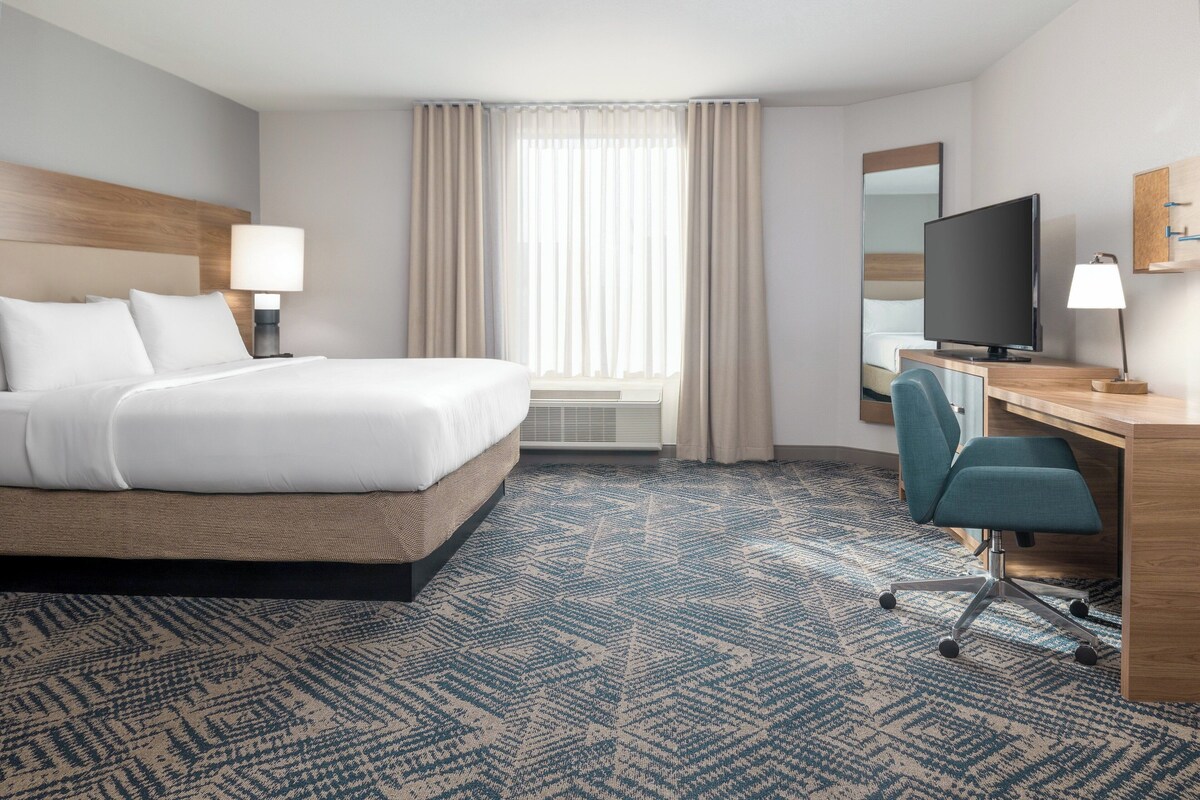 A hotel room with a bed, desk, chair, television, and a large window with curtains.
