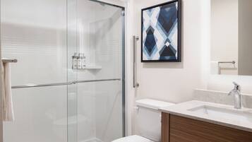 A bathroom with a glass shower enclosure, a white toilet, a sink with a marble countertop, and a framed abstract artwork on the wall.