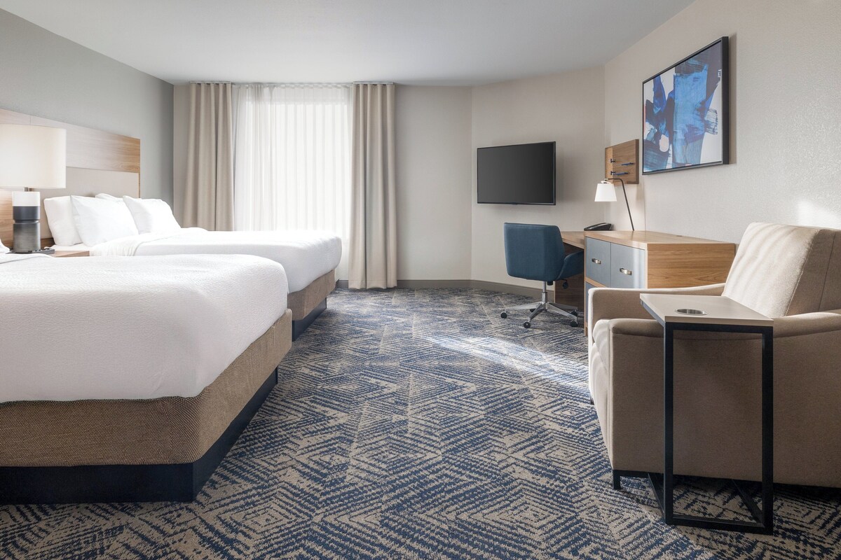A hotel room with two beds, a desk, a chair, a television, and a large window with curtains.