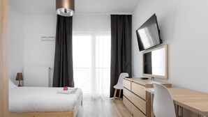 A modern bedroom with a bed, a desk, a chair, a TV, and a window with curtains. - Wola Chic Brown Studio (Warsaw)