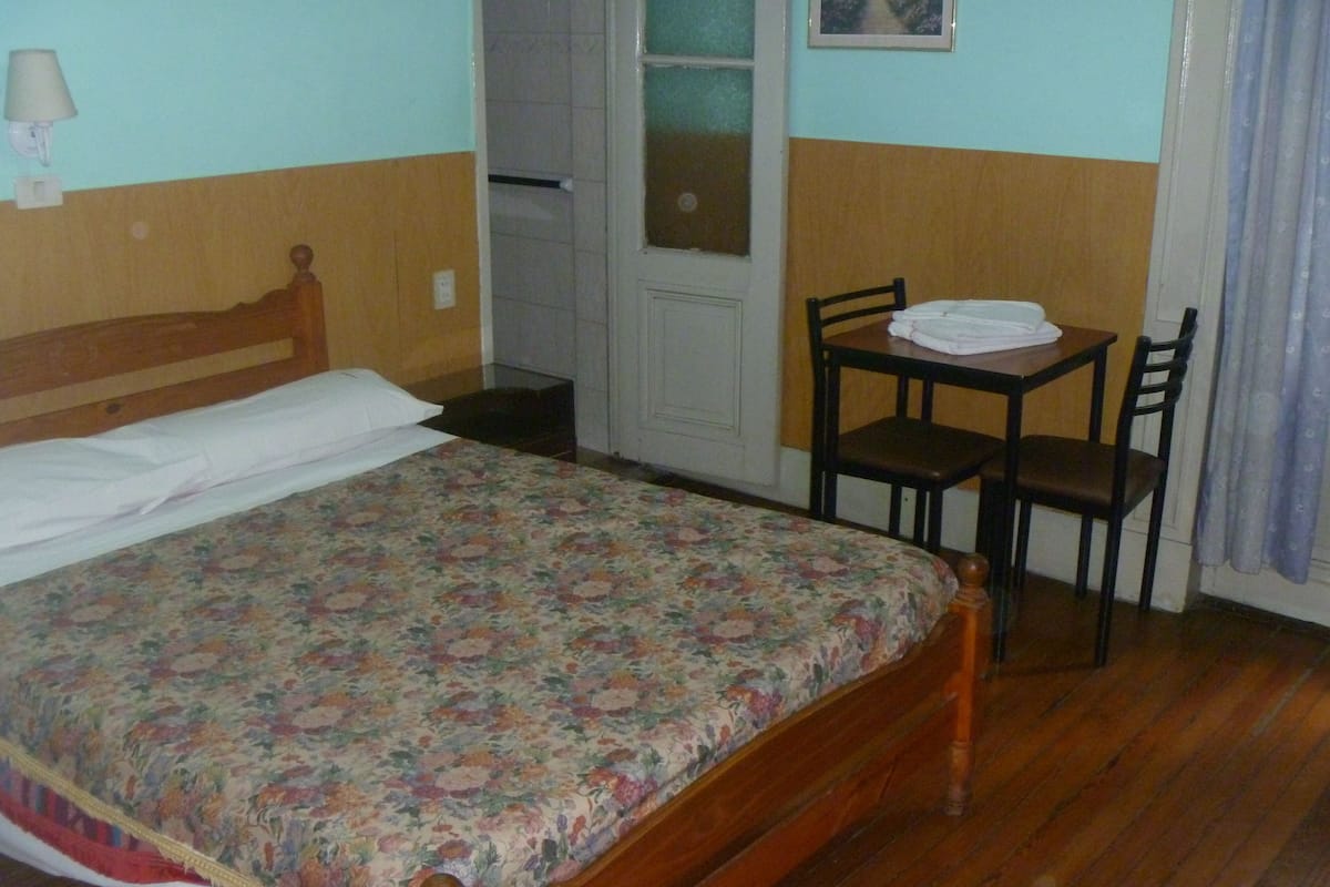A room with a wooden bed, a small table with chairs, a window with a view, and a door.