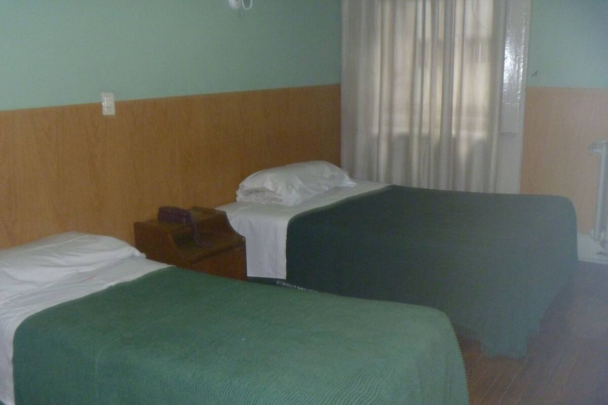 A room with two single beds, a wooden nightstand, a white wall, and a window with curtains.