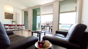 A modern living room with a sofa, a coffee table with colorful balls, a dining area with chairs, and a view of the sea through large windows.