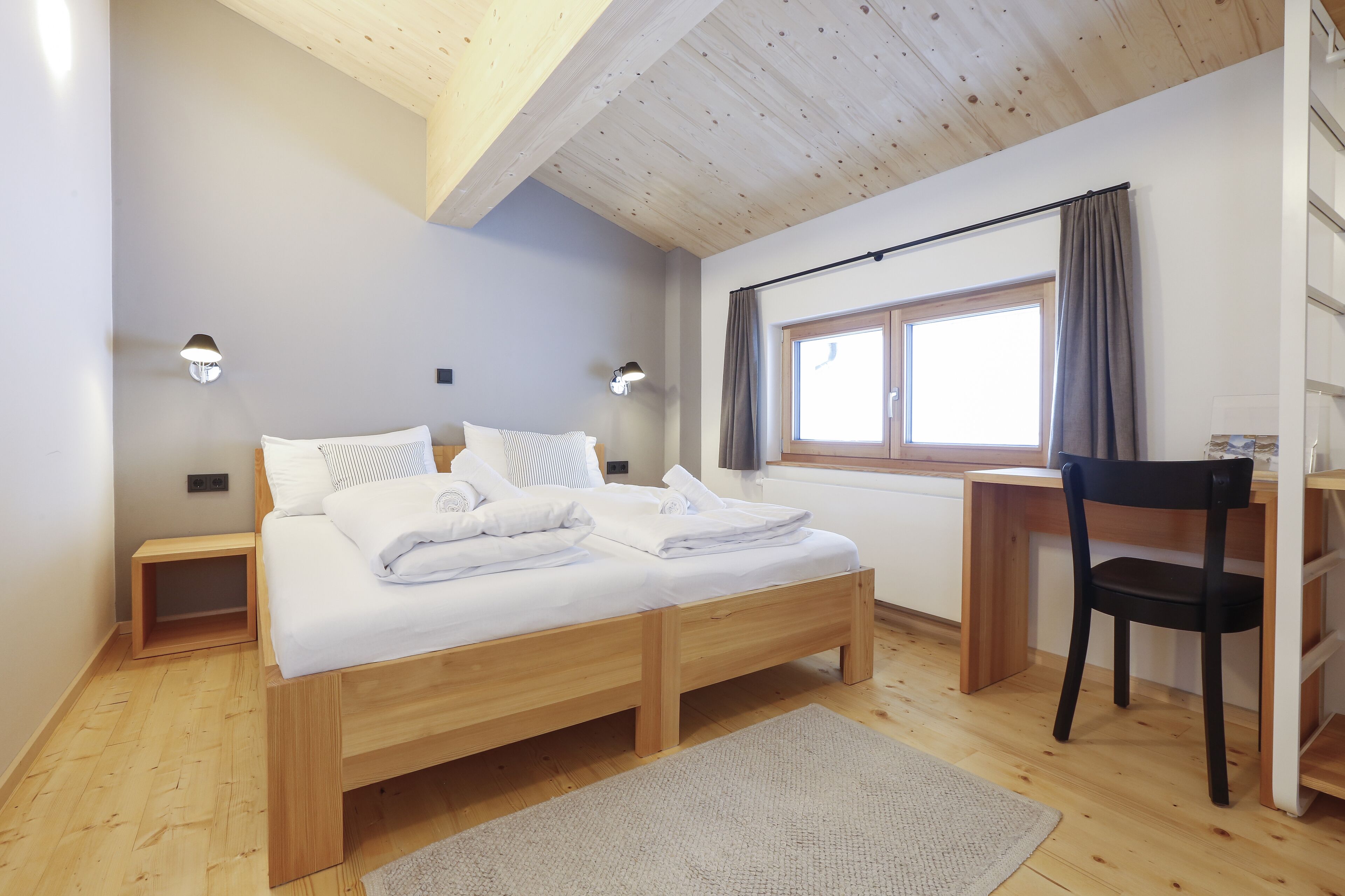A bedroom with a wooden bed, bedside tables, a desk, a chair, and a window with curtains.