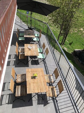 Eine Außenpatio mit mehreren Holztischen und Stühlen, ein überdachter Bereich mit Sonnenschirm und eine Brüstung mit Blick auf eine Rasenfläche.