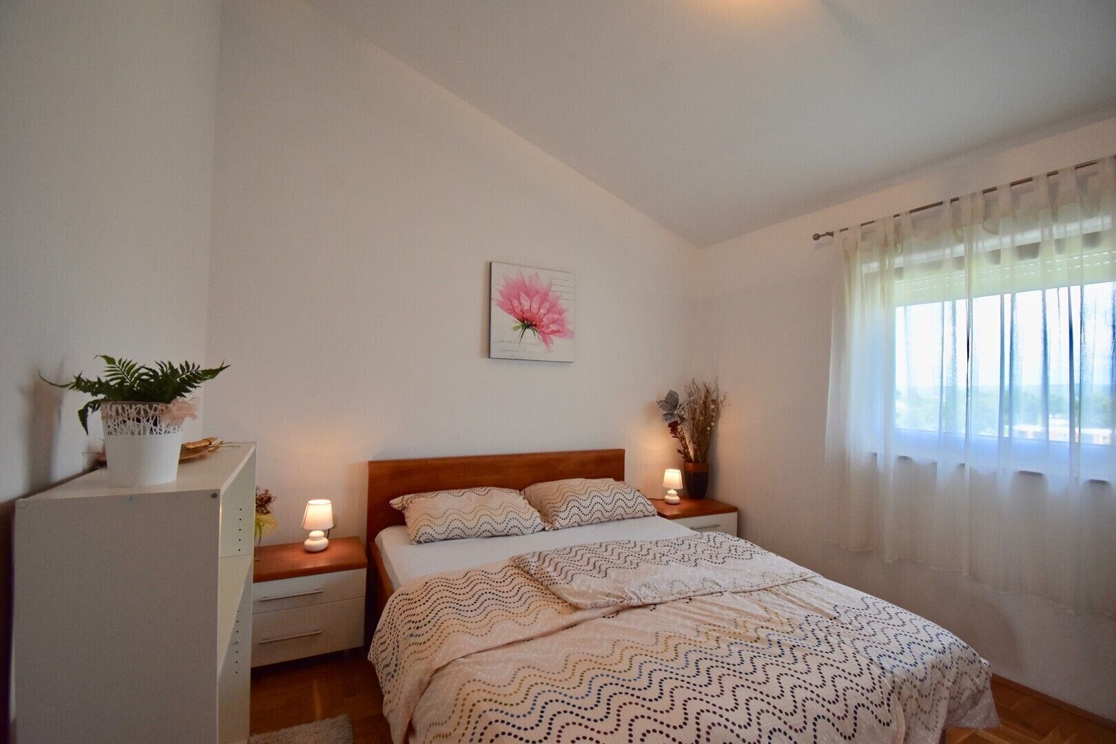 Quarto com cama, mesas de cabeceira, planta em vaso, quadro na parede e janela com cortinas leves.