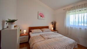 A bedroom with a bed, nightstands, a potted plant, a wall-mounted painting, and a window with sheer curtains. - Comfortable apartment near the Hidrobaza beach (Pula)