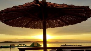A beach umbrella with a thatched roof, overlooking a sunset over the ocean with boats and rocks in the distance. - Comfortable apartment near the Hidrobaza beach (Pula)