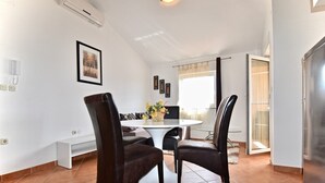 A dining area with a round table, two chairs, and a vase with flowers. There is a window with curtains, a door, and a wall-mounted air conditioner. - Comfortable apartment near the Hidrobaza beach (Pula)