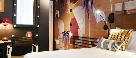 Habitación de hotel con cama, escritorio y silla, espejo con luces, y pared con diseño decorativo.