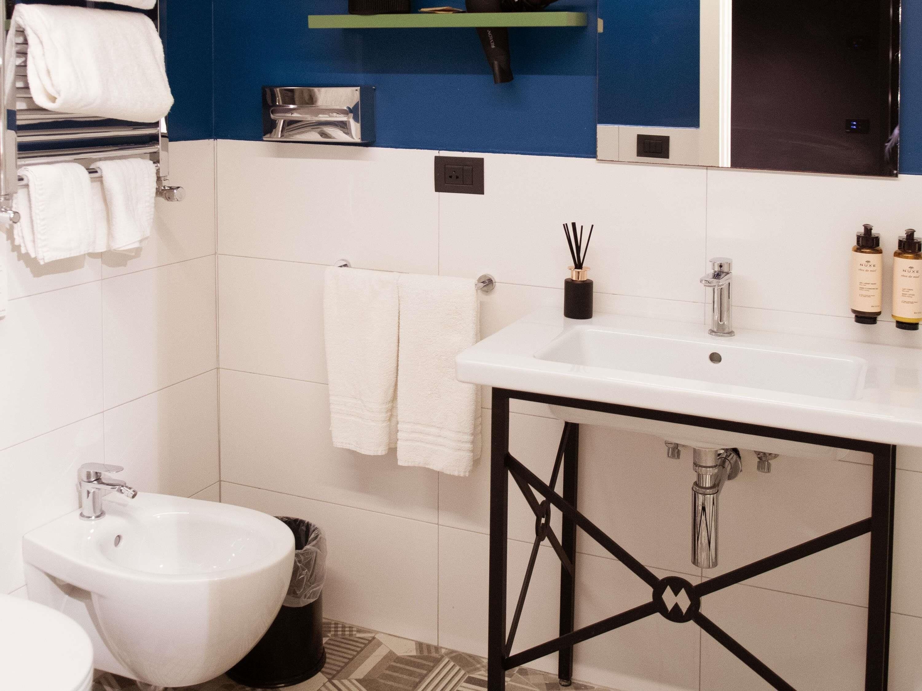 Ein modernes Badezimmer mit einem weißen Waschbecken, einem schwarzen Metallgestell und einem Bidet. Weiße Handtücher hängen an einem Halter, ein kleines Regal mit Toilettenartikeln und ein Spiegel über dem Waschbecken.