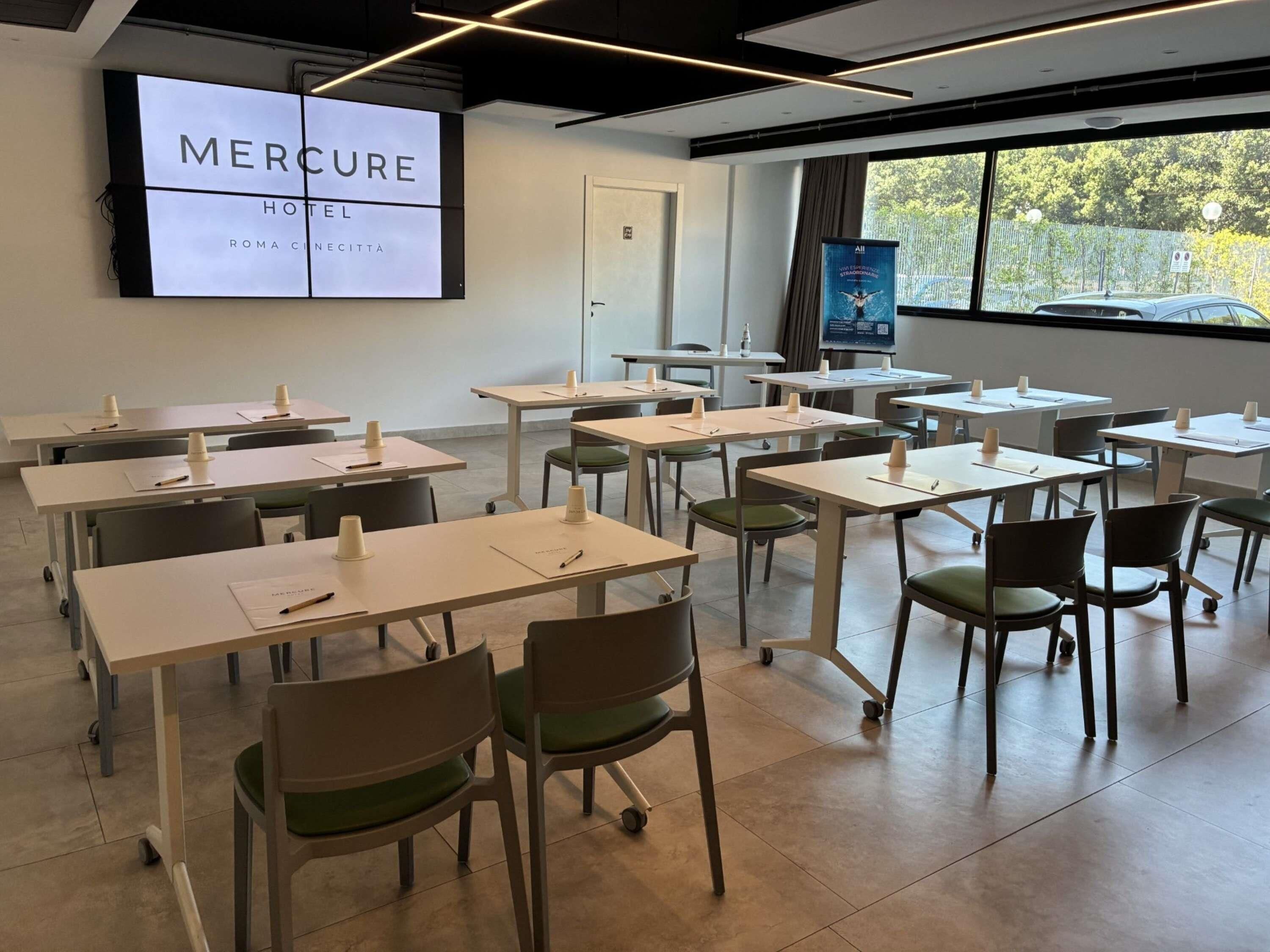 Una sala de conferències amb taules, cadires i una pantalla gran que mostra 'MERCURE HOTEL ROMA CINCETTA'.