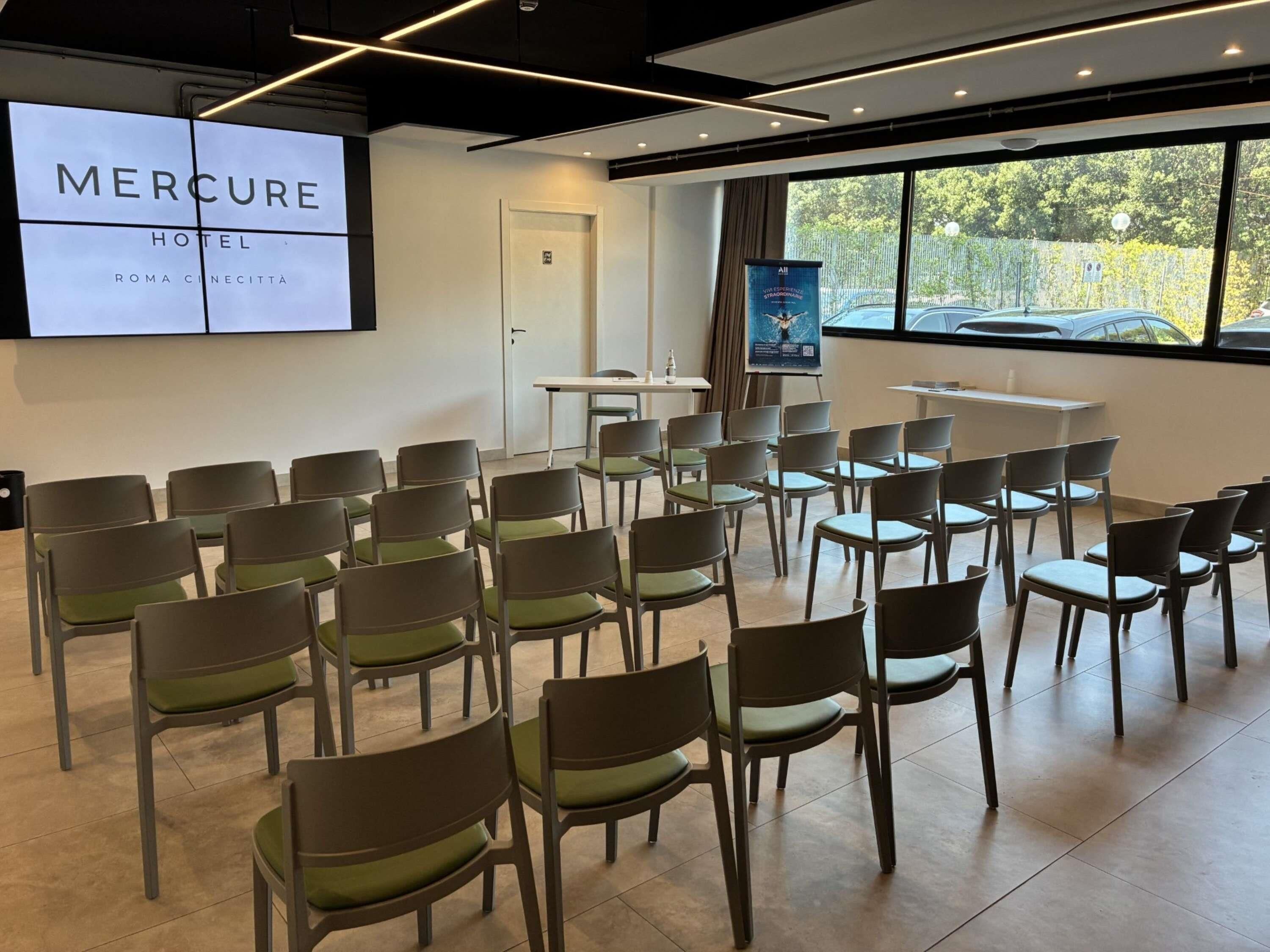 Un salón de conferencias con filas de sillas, una pantalla de proyector mostrando 'MERCURE HOTEL ROMA CINCETTA' y un cartel en la pared.