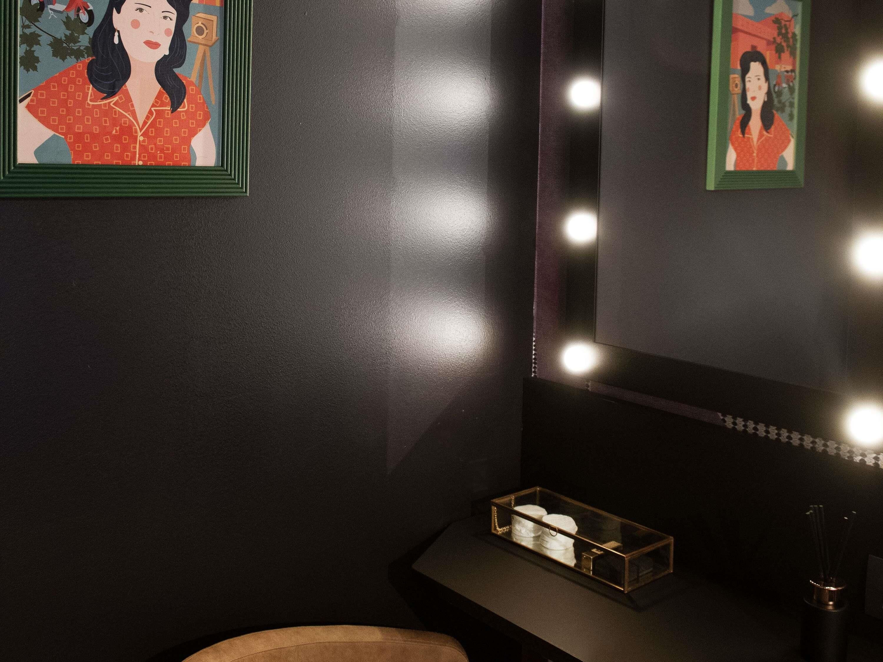 A room with a dark wall, a mirror with lights, and two framed pictures.