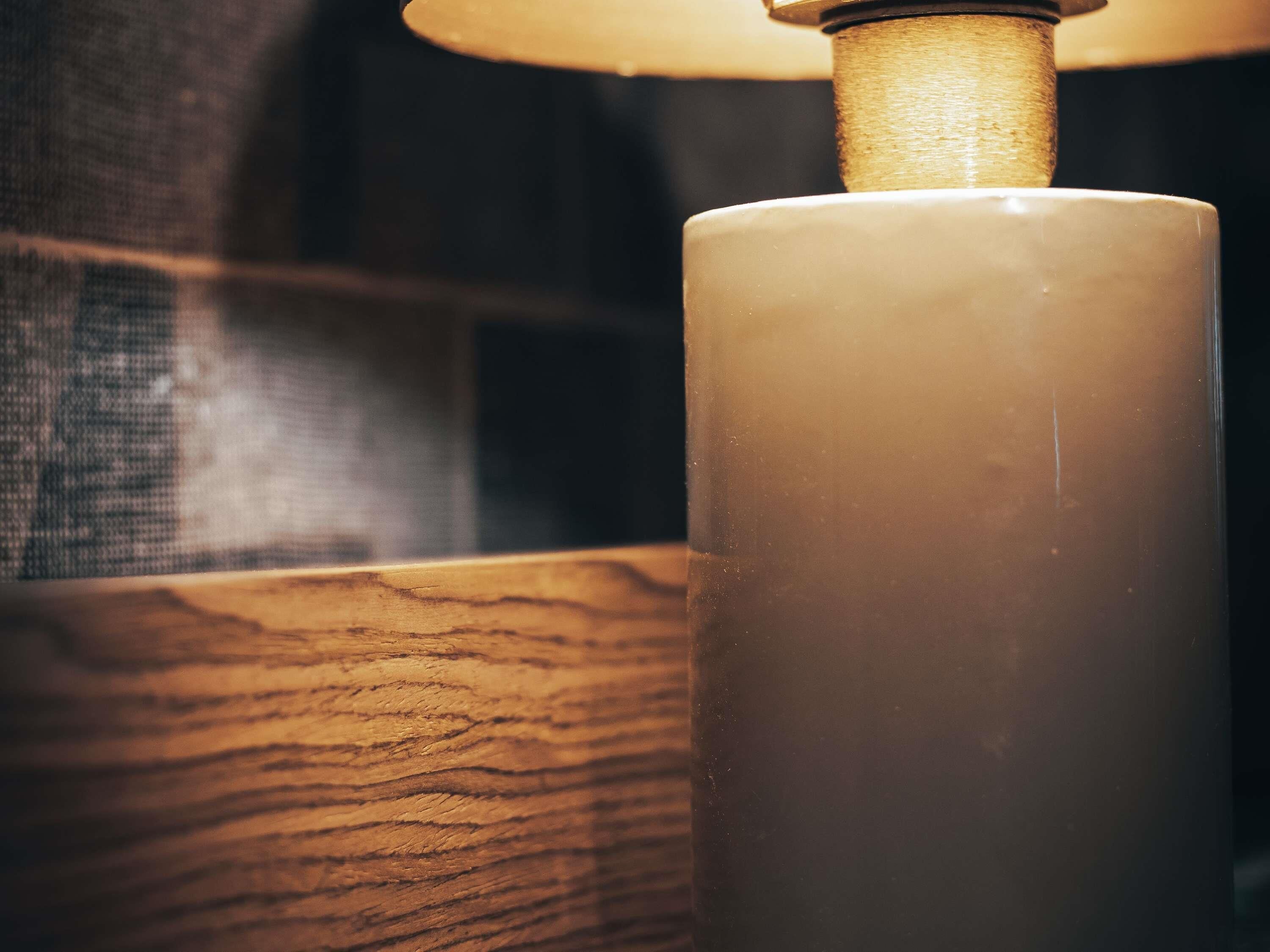 A close-up of a white candle in a glass holder with a wooden base, set against a blurred background.