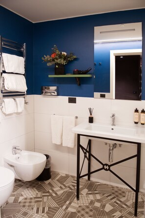 A bathroom with a white sink, black metal frame, white tiles, a white toilet, a bidet, a trash can, a towel rack, a mirror, and a shelf with toiletries. - Mercure Roma Cinecittà  (Rome)