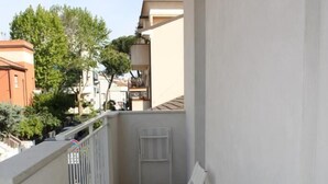 Un pequeño balcón con una mesa de vidrio y dos sillas blancas, barandilla blanca y vistas a edificios y árboles vecinos.