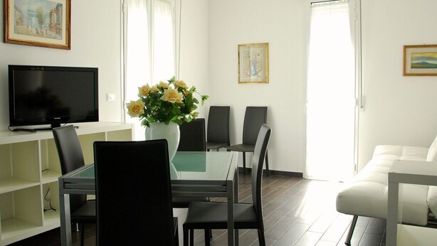 Una sala de estar moderna con una mesa de comedor de vidrio, sillas negras, un sofá blanco, un televisor de pantalla plana y un jarrón con flores.
