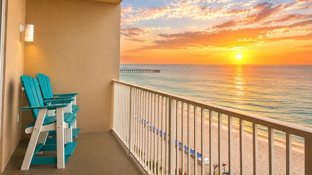A balcony with a view of the sunset over the ocean, a pier, and beach umbrellas. - Amazing View - 6th Floor Gulf Front Condo - Close to Pier Park (Panama City Beach)