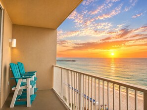 Een balkon met uitzicht op de ondergaande zon boven de oceaan, een pier en strandparasols.