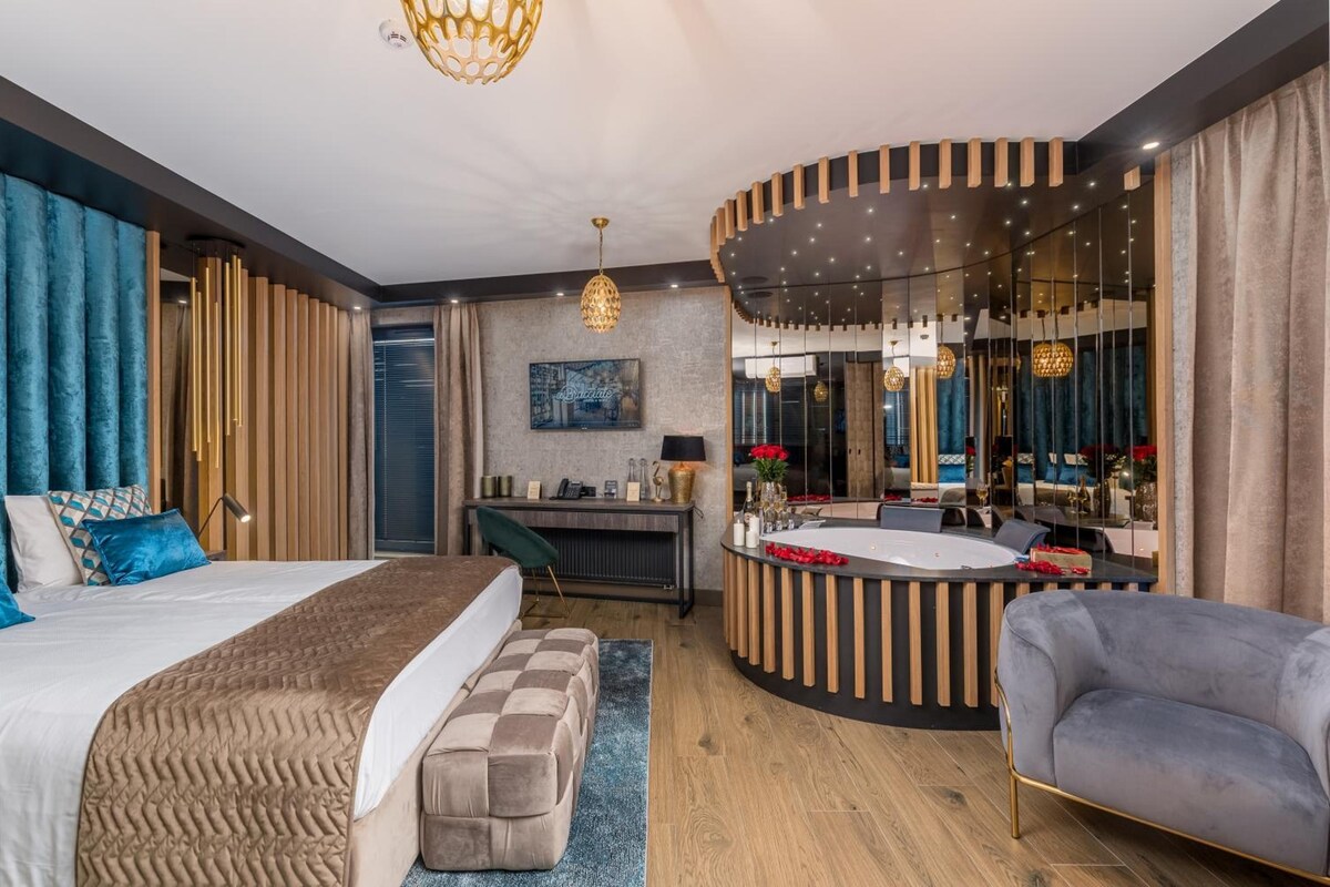 Una habitación de hotel moderna con una cama grande, una zona de estar con escritorio, un baño con bañera y un espejo grande.