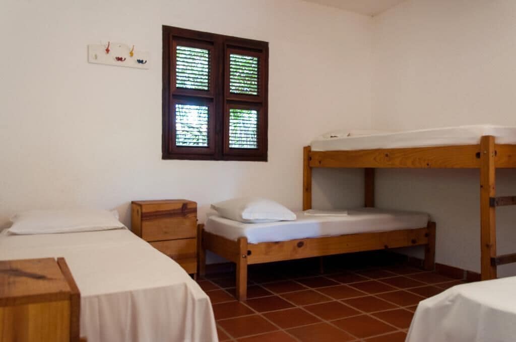 A room with two single beds, a wooden nightstand, a window with blinds, and a tiled floor.