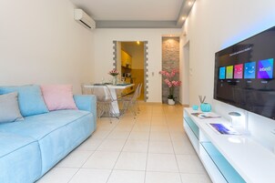 Una sala de estar moderna con un sofá azul, un comedor con mesa y sillas, un televisor de pantalla plana montado en la pared y una cocina visible a través de un vano abierto.