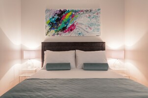 A neatly made bed with a white and teal color scheme, a modern headboard, and a colorful abstract painting above it.