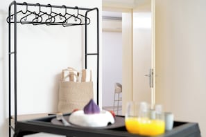 Una habitación con un perchero metálico, una cesta tejida, una bandeja con una almohada, vasos y una botella de jugo de naranja.