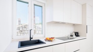 A modern kitchen with a black sink, white cabinets, a window with a view of buildings, and a countertop with a coffee maker and fruit. - Apartments Stipisic (Hvar)