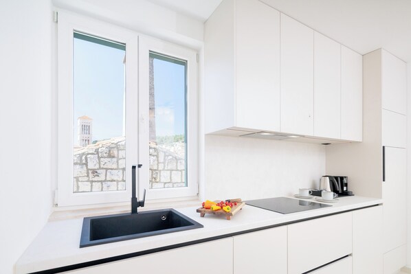 Eine moderne Küche mit einem schwarzen Spülbecken, weißen Schränken, einem Fenster mit Blick auf Gebäude und einer Arbeitsplatte mit Kaffeemaschine und Obst.