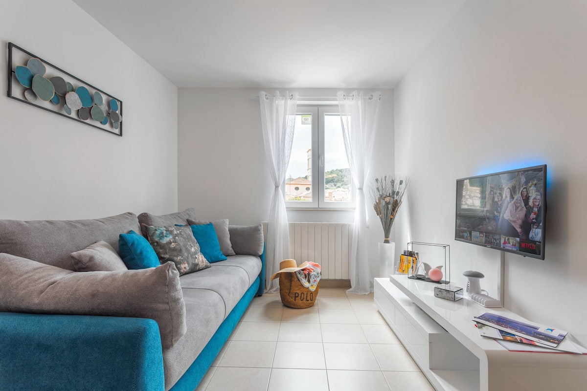 A modern living room with a grey sofa, a basket, a TV, and a window with curtains.
