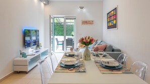 A modern dining area with a table set for four, clear chairs, a glass-top console table, and a television on the left. - Apartments Stipisic (Hvar)