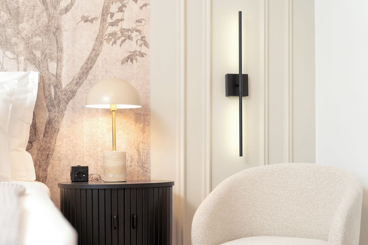A modern room with a round side table, a lamp, a wall-mounted light, a chair, and a wall with a tree mural.