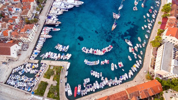 An aerial view of a marina with numerous boats docked in a circular pattern, surrounded by buildings with red-tiled roofs. - Apartments Stipisic (Hvar)