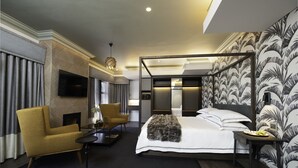Une chambre d’hôtel moderne dotée d’un grand lit, d’un coin salon avec un fauteuil et une table, d’un téléviseur à écran plat et d’un mur décoratif orné d’un motif de feuilles.